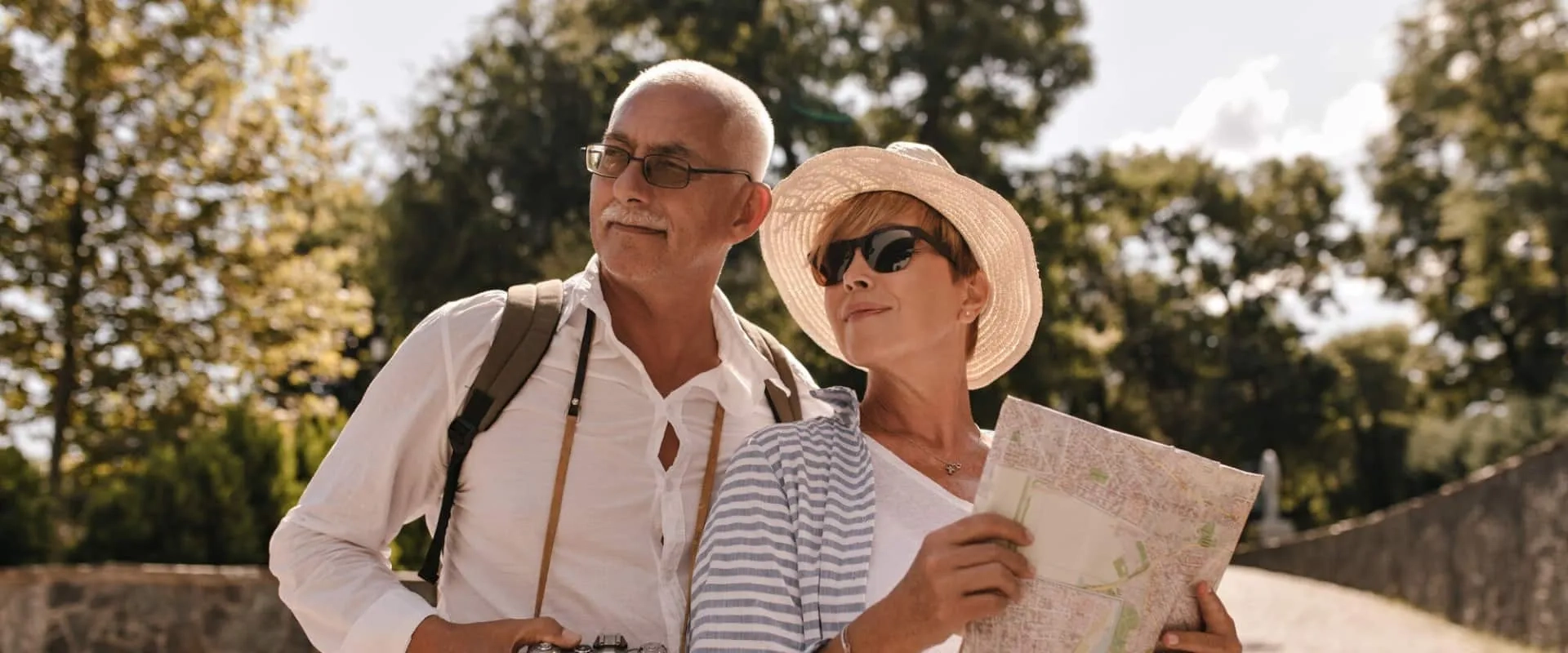 Una pareja de adultos mayores, el hombre con cabello canoso y gafas, y la mujer con un sombrero y gafas de sol, están de pie al aire libre.