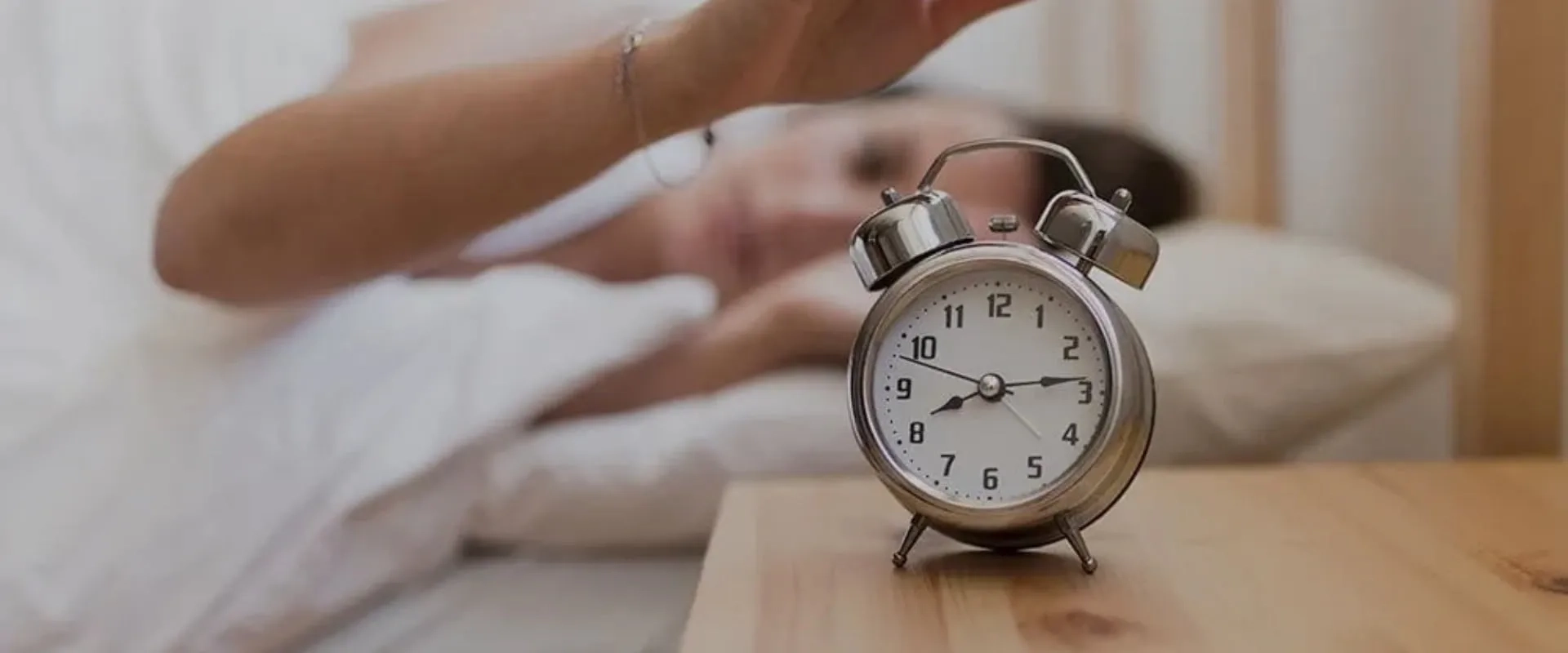 Una persona en la cama intenta apagar un despertador antiguo que suena. El reloj, de metal plateado, está en una mesa de noche de madera y muestra la hora.