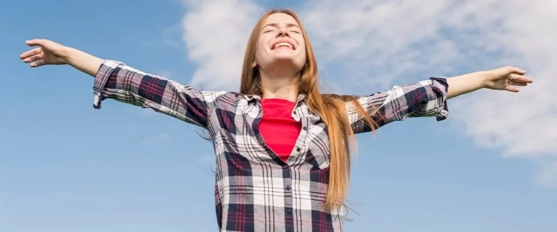Una joven sonriente con el cabello largo y suelto está de pie bajo un cielo azul despejado, con los brazos extendidos.
