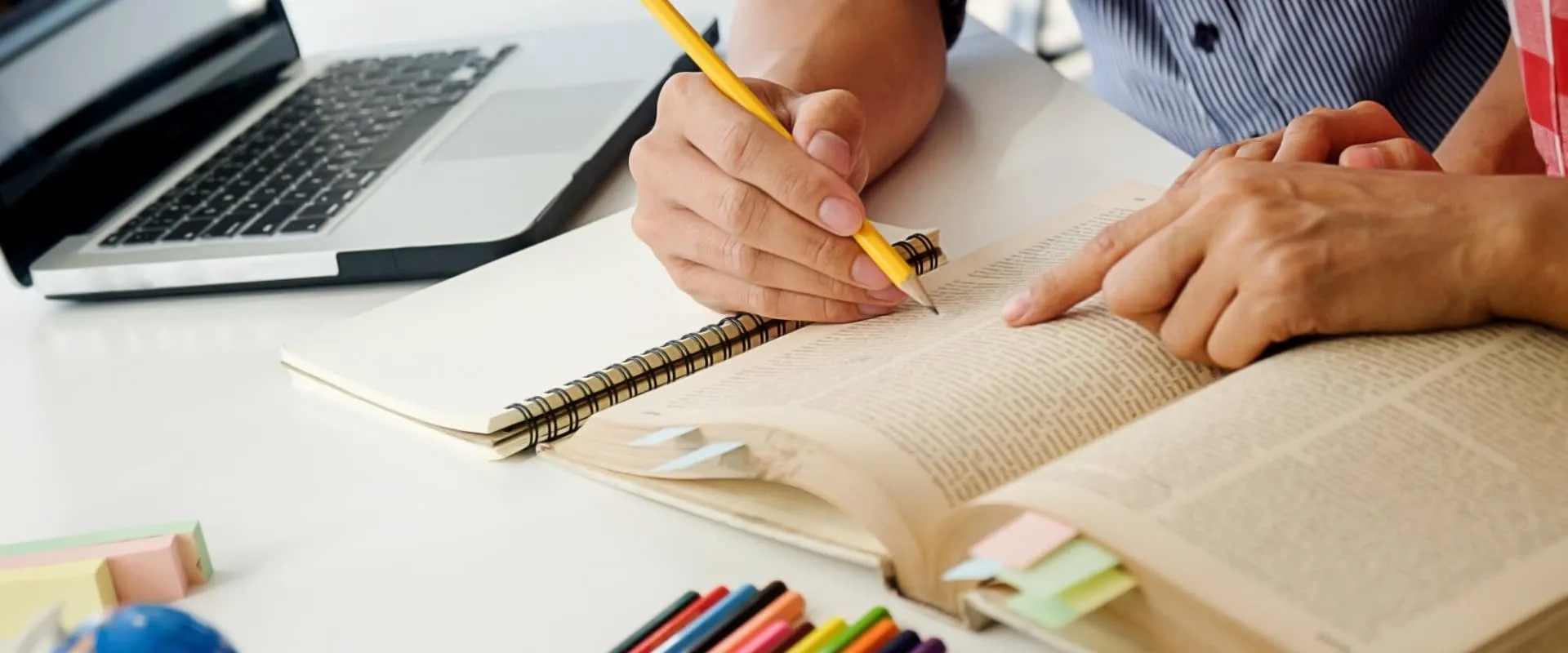 Dos manos trabajan juntas sobre un libro abierto, con una de ellas señalando un texto mientras la otra sostiene un lápiz. Junto al libro, hay un cuaderno, lápices de colores y una laptop visible, creando un ambiente de estudio colaborativo.