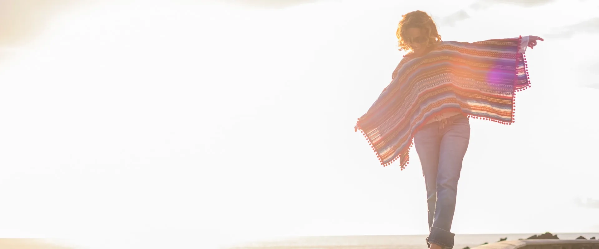 Una mujer caminando sobre un muro de piedra al borde del mar, vistiendo un poncho de rayas coloridas. 