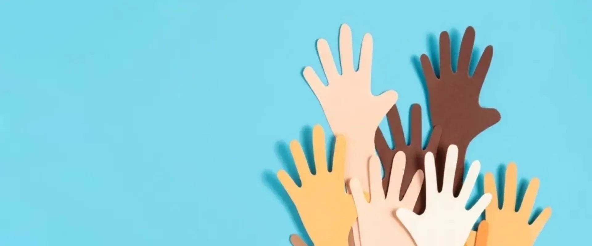 Un conjunto de manos de diferentes tonos de piel se levantan sobre un fondo azul claro. Las manos son de papel recortado, simbolizando diversidad y unidad.