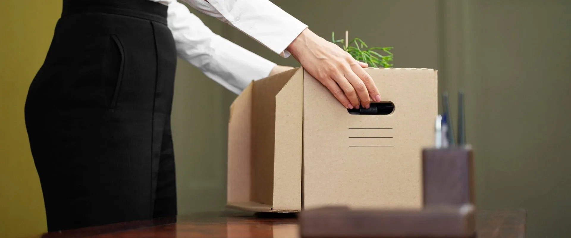 Un primer plano de una mano femenina colocando un objeto en una caja de cartón, que contiene algunas plantas y artículos de oficina. 