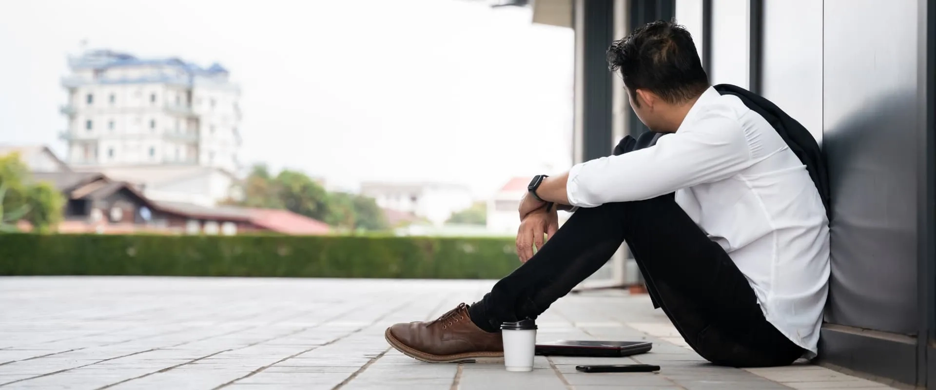 Un hombre sentado en el suelo, de espaldas, con una expresión pensativa. Lleva una camisa blanca y pantalones oscuros, y a su lado hay un portátil y un vaso de café. 