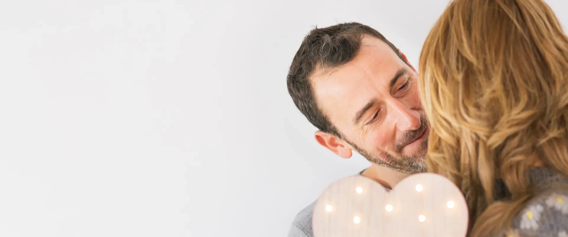 Un hombre sonriente mira cariñosamente a una mujer, mientras ambos están en un entorno iluminado y acogedor. La mujer tiene el cabello largo y ondulado, y ambos parecen disfrutar de un momento íntimo. 