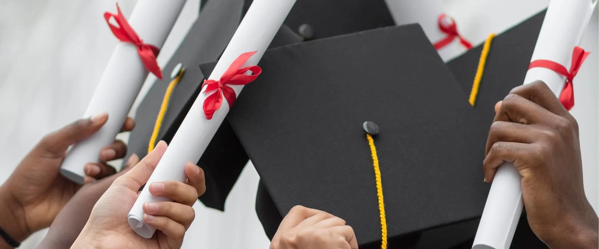 Un grupo de manos sostiene diplomas enrollados con cintas rojas, celebrando una graduación. Los diplomas son sostenidos junto a birretes de graduación negros, creando una atmósfera de logro y celebración. 