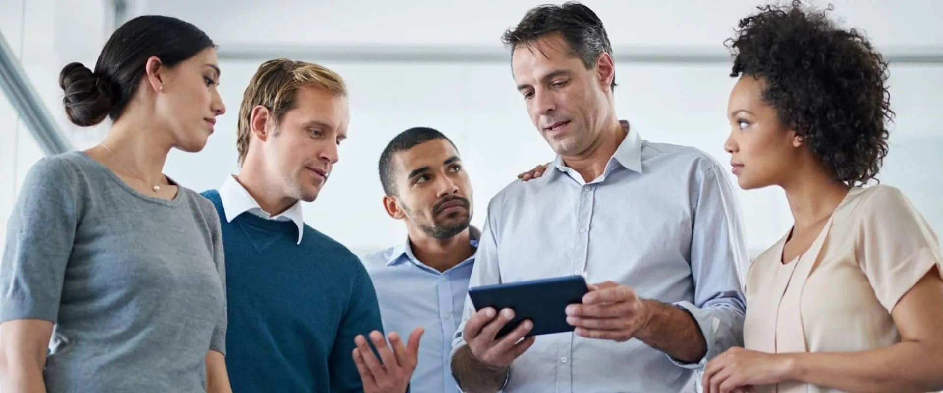 Cinco personas adultas, hombres y mujeres, observan atentamente una tablet mientras conversan en un entorno laboral; uno de ellos sostiene el dispositivo mientras los demás lo rodean con actitud de colaboración.