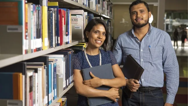 Dos estudiantes o profesionales de posgrado, un hombre y una mujer, sonriendo y sosteniendo libros y carpetas. Están de pie en el pasillo de una biblioteca, con estanterías llenas de libros a un lado, simbolizando la investigación académica y el estudio de posgrado.